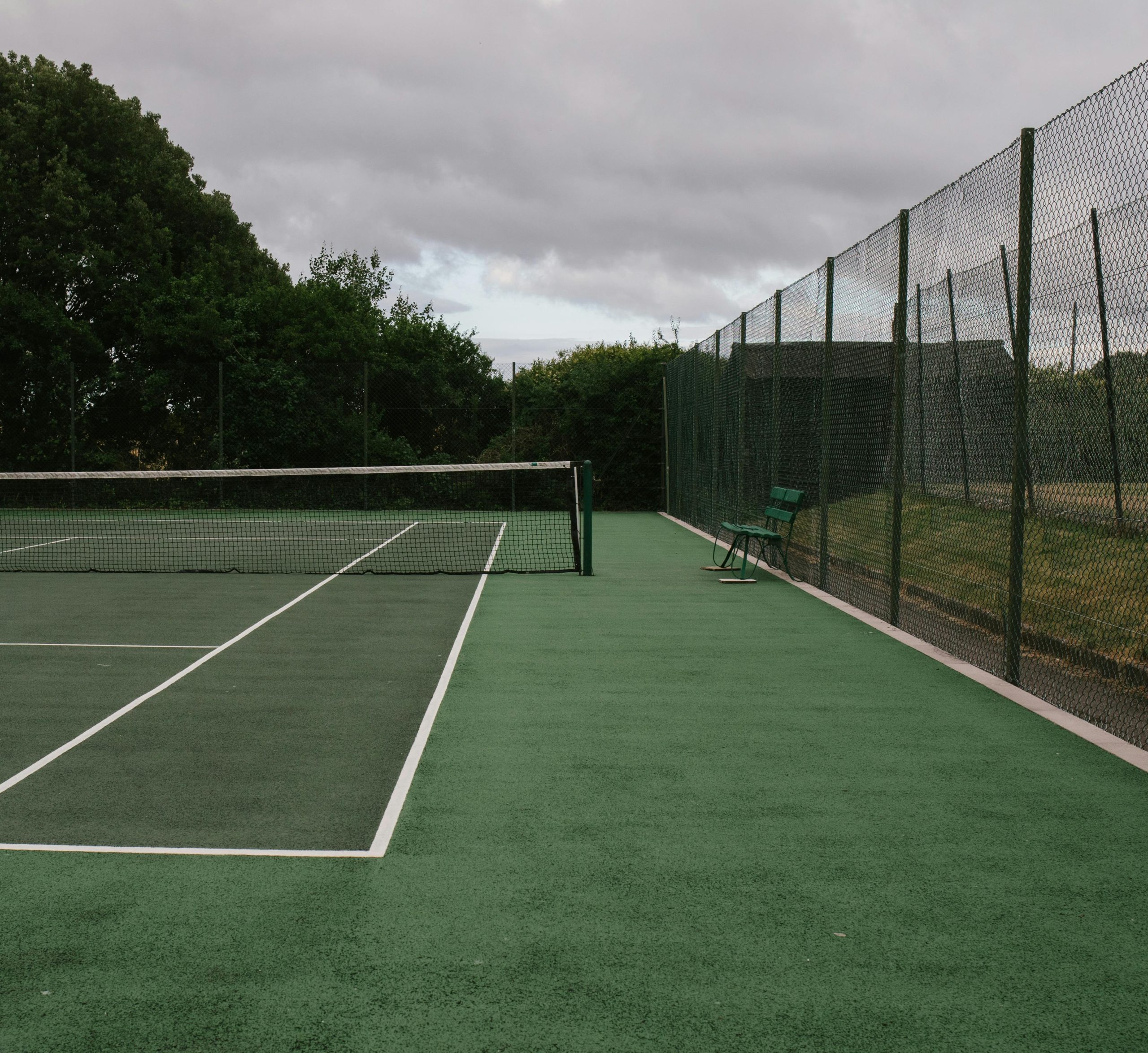 Green tennis court with net and fence under a cloudy sky, ideal for sports concepts.